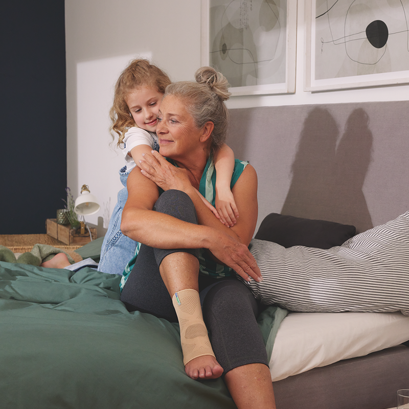 Woman wearing an ankle support sits on bed in bedroom while small girl hugs her from behind 